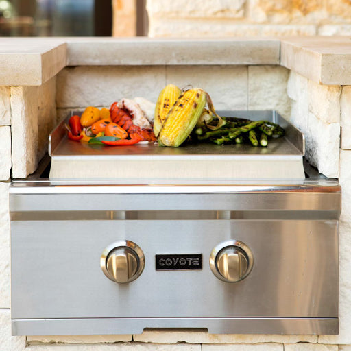 Stono Outdoor Living The Coyote Teppanyaki Plate - CTEP by Coyote is a stainless steel outdoor grill with a flat surface, built into a stone countertop, featuring two control knobs and showcasing grilled corn, asparagus, bell peppers, and lobster tails.