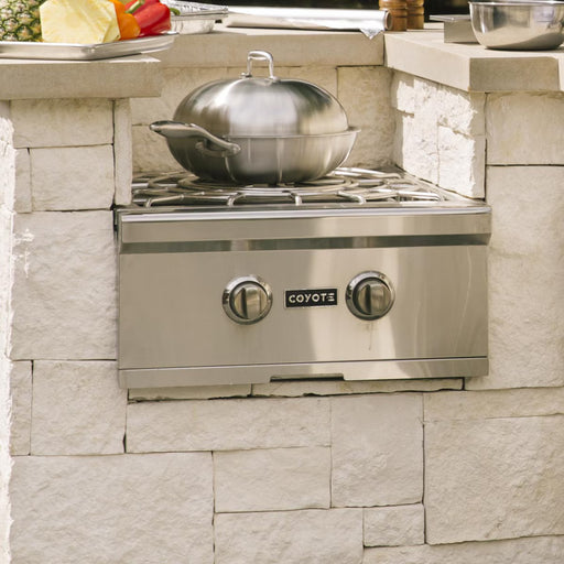 Stono Outdoor Living A Coyote Wok for Power Burner (CWOK) by Coyote sits atop a stainless steel outdoor gas stove with two knobs, built into a stone counter. A covered pot, fresh vegetables, and a cutting board suggest an upcoming stir-fry.