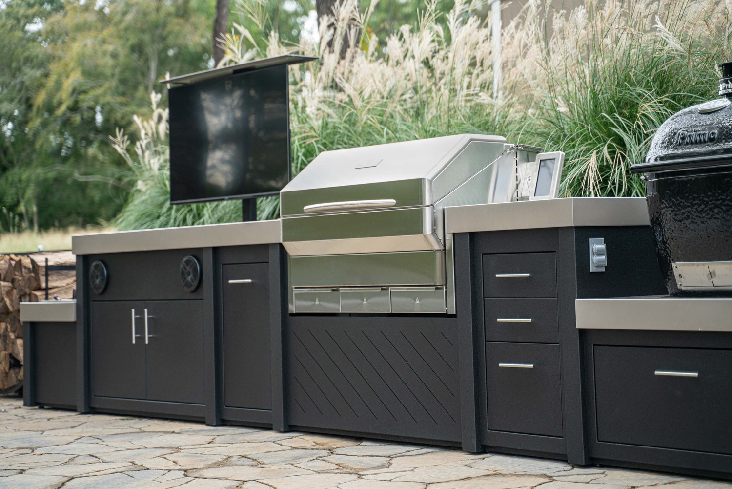 Stono Outdoor Living A modern outdoor kitchen featuring a stainless steel grill, black cabinets with silver handles, a mounted TV, and a ceramic smoker, set on a stone patio with tall ornamental grasses behind.