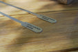 Stono Outdoor Living Two Tagwood BBQ Set of 2 Stainless Steel Skewers (BBQ89) with flat engraved ends rest on a wooden surface, sunlight casting shadows—perfect for your next portable BBQ.