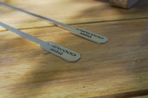 Stono Outdoor Living Two Tagwood BBQ Set of 2 Stainless Steel Skewers (BBQ89) with flat engraved ends rest on a wooden surface, sunlight casting shadows—perfect for your next portable BBQ.
