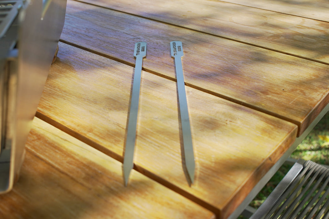 Stono Outdoor Living Two Tagwood BBQ Set of 2 Stainless Steel Skewers (model BBQ89) rest parallel on a wooden outdoor table, with dappled sunlight and partial shadows across the surface.