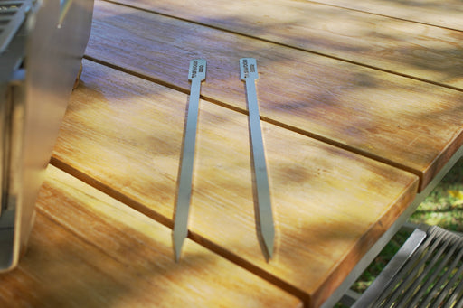 Stono Outdoor Living Two Tagwood BBQ Set of 2 Stainless Steel Skewers (model BBQ89) rest parallel on a wooden outdoor table, with dappled sunlight and partial shadows across the surface.