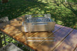 Stono Outdoor Living A Tagwood BBQ portable stainless steel grill sits on a wooden table, with a Tagwood BBQ Set of 2 Stainless Steel Skewers (BBQ89) in front. A grassy area and garden chair make it perfect for outdoor grilling with these durable skewers.