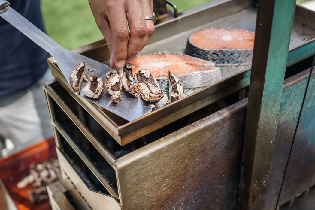 Stono Outdoor Living A hand places sliced mushrooms onto a Tagwood BBQ Height Adjustable Griddle - BBQ54SS, a 304 stainless steel extension by Tagwood BBQ, next to raw salmon steaks on an outdoor grill.