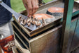 Stono Outdoor Living A hand places sliced mushrooms onto a Tagwood BBQ Height Adjustable Griddle - BBQ54SS, a 304 stainless steel extension by Tagwood BBQ, next to raw salmon steaks on an outdoor grill.