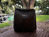 Stono Outdoor Living A Tagwood BBQ Grill Cover for BBQ23SS Grill - BBQ86SS by Tagwood BBQ protects a large grill on a wooden deck in a green backyard near trees and a canal with a boat.