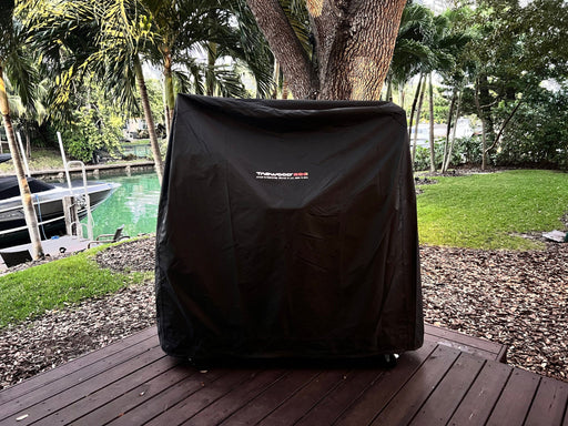 Stono Outdoor Living A Tagwood BBQ Grill Cover for BBQ23SS Grill - BBQ86SS by Tagwood BBQ protects a large grill on a wooden deck in a green backyard near trees and a canal with a boat.