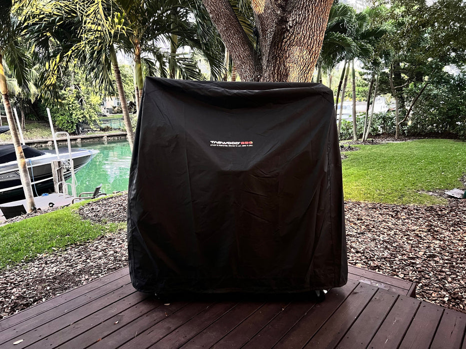 Stono Outdoor Living A Tagwood BBQ Grill Cover for BBQ23SS Grill - BBQ86SS by Tagwood BBQ protects a large grill on a wooden deck in a green backyard near trees and a canal with a boat.