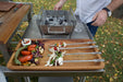 Stono Outdoor Living A person stands by a grill with a Tagwood BBQ Set of 2 Stainless Steel Skewers (BBQ89) loaded with colorful veggies and a sauce bowl on a wooden tray. The grill has hot coals, and the table sits outdoors on grass with autumn leaves.