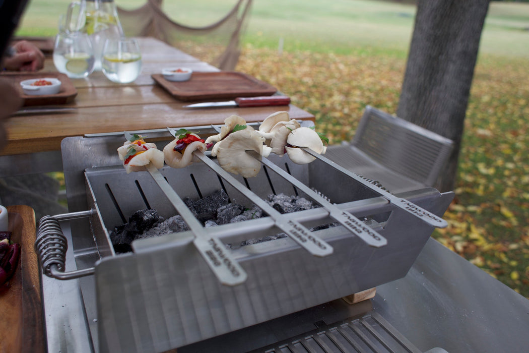 Stono Outdoor Living Tagwood BBQ Set of 2 Stainless Steel Skewers (BBQ89) are grilling vegetables and mushrooms over charcoal in a rectangular tabletop brazier, with drinks and plates on a wooden table outdoors.