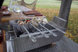 Stono Outdoor Living Tagwood BBQ Set of 2 Stainless Steel Skewers (BBQ89) are grilling vegetables and mushrooms over charcoal in a rectangular tabletop brazier, with drinks and plates on a wooden table outdoors.