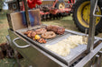 Stono Outdoor Living The Tagwood BBQ Interchangeable Griddle for BBQ01SS (BBQ57-01SS) cooks chicken, sausages, skewers, cabbage, and more. Red bell peppers hang above; a tractor wheel and farm equipment appear in the background. Ideal for shrimp too!.