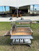 Stono Outdoor Living The Tagwood BBQ Round Warming Roof Grill for BBQ23SS/BBQ25SS - BBQ99SS, featuring 304 stainless steel racks and ample cooking space, is set up on grass in front of an empty stage with rows of chairs and a palm tree under a white canopy.