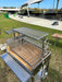 Stono Outdoor Living The Tagwood BBQ Round Warming Roof Grill for BBQ03SS/BBQ05SS - BBQ92SS by Tagwood BBQ, made of 304 stainless steel, features multiple cooking racks and a side wheel, set up outdoors near a pavilion with black chairs and tables.