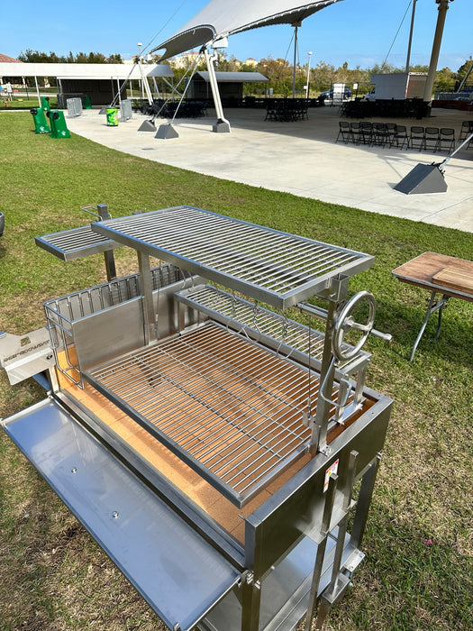 Stono Outdoor Living The Tagwood BBQ Round Warming Roof Grill for BBQ23SS/BBQ25SS - BBQ99SS by Tagwood BBQ, made of 304 stainless steel, with adjustable grates and warming rack, is set up on grass near a wooden table and pavilion with chairs under a white canopy.