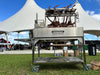 Stono Outdoor Living The Tagwood BBQ Round Warming Roof Grill for BBQ03SS/BBQ05SS - BBQ92SS is shown outdoors on grass in front of a white tent beneath a blue sky, with people and event booths visible in the background. Tomahawk steaks cook on its 304 stainless steel rack.
