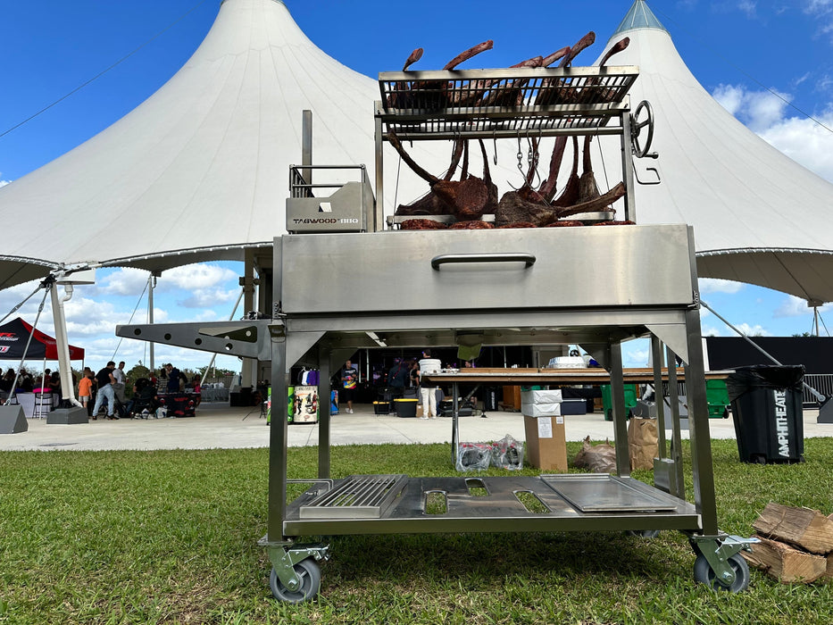 Stono Outdoor Living The Tagwood BBQ Round Warming Roof Grill for BBQ23SS/BBQ25SS - BBQ99SS is set up on the grass, with event tables and guests under a modern white tent against a blue sky. Brand: Tagwood BBQ.