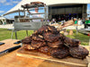 Stono Outdoor Living A large stack of grilled steaks and tomahawk ribeyes on a wooden board outdoors, with the spacious Tagwood BBQ Round Warming Roof Grill for BBQ09SS - BBQ98SS by Tagwood BBQ, a person, and a big tent visible on a sunny day.