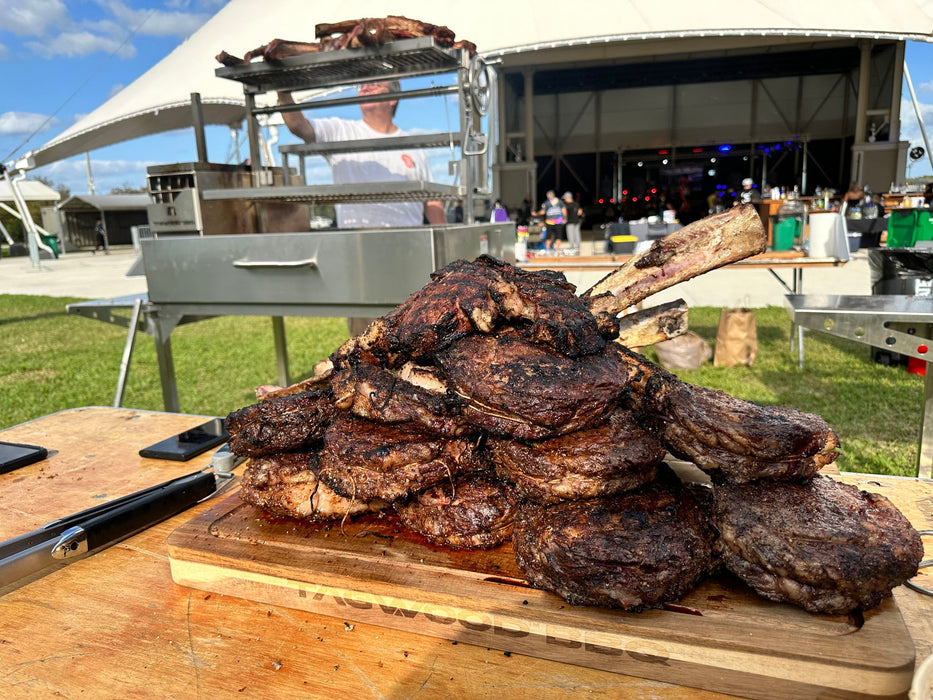 Stono Outdoor Living A stack of grilled steaks and chops on a cutting board sits beside the Tagwood BBQ Round Warming Roof Grill for BBQ23SS/BBQ25SS - BBQ99SS, featuring a 304 stainless steel warming rack, as someone cooks in the background at an outdoor event.