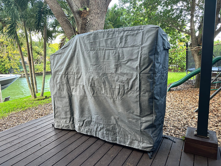 Stono Outdoor Living A large grill with a Tagwood BBQ BBQ26SS Cover - COVE26 from Tagwood BBQ sits on a wooden deck near a tree, with a canal, lawn, and playground slide in the background.
