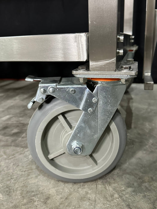Stono Outdoor Living Close-up of a metal swivel caster with brake, attached to the leg of the Tagwood BBQ Argentine Santa Maria Wood Fire & Charcoal Grill - BBQ26SS by Tagwood BBQ, resting on a concrete floor.