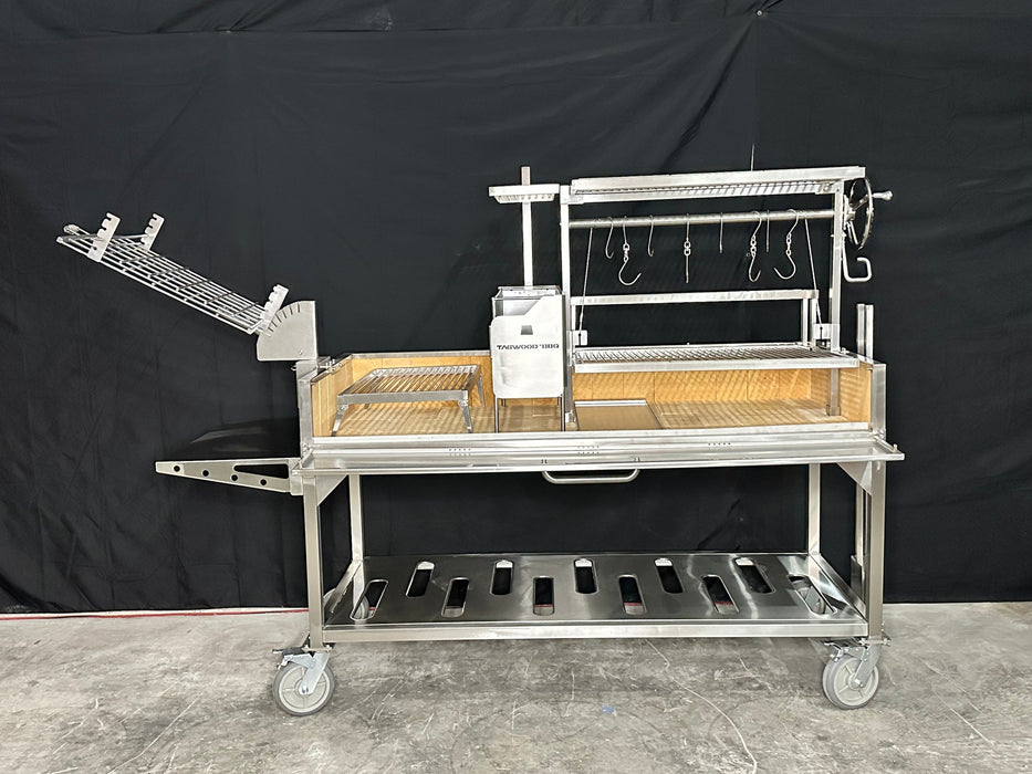 Stono Outdoor Living The Tagwood BBQ Argentine Santa Maria Wood Fire & Charcoal Grill (BBQ26SS) features 304 stainless steel, wood accents, wheels, grilling racks, hooks, a side basket, and a handle—classic Argentine design—shown on a plain black background.