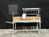 Stono Outdoor Living The Tagwood BBQ Argentine Santa Maria Wood Fire & Charcoal Grill - BBQ26SS features 304 stainless steel, a rotisserie, grilling hooks, mesh rack, crank handle, and lower tray—shown on concrete flooring with a black backdrop.