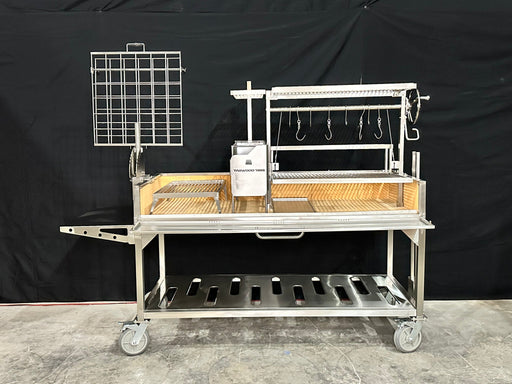 Stono Outdoor Living The Tagwood BBQ Argentine Santa Maria Wood Fire & Charcoal Grill - BBQ26SS features 304 stainless steel, a rotisserie, grilling hooks, mesh rack, crank handle, and lower tray—shown on concrete flooring with a black backdrop.