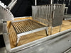 Stono Outdoor Living A close-up of the Tagwood BBQ Argentine Santa Maria Wood Fire & Charcoal Grill – BBQ26SS, made from 304 stainless steel with a removable grilling rack and vertical meat racks, placed on a tiled base against a dark background.