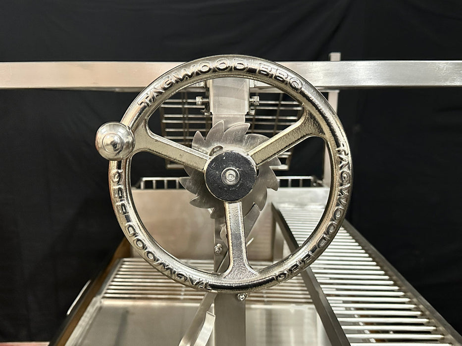 Stono Outdoor Living Close-up of a metal hand crank engraved with Tagwood BBQ, attached to the Tagwood BBQ Argentine Santa Maria Wood Fire & Charcoal Grill - BBQ26SS, with stainless steel grates visible against a black background.