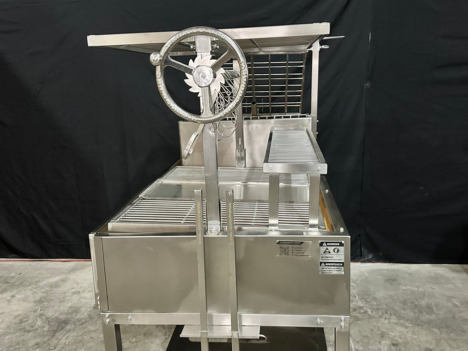 Stono Outdoor Living The Tagwood BBQ Argentine Santa Maria Wood Fire & Charcoal Grill - BBQ26SS by Tagwood BBQ is a large 304 stainless steel grill with a hand crank wheel, metal grates, black backdrop, and warning labels on the front panel.
