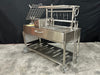 Stono Outdoor Living The Tagwood BBQ Argentine Santa Maria Wood Fire & Charcoal Grill (BBQ26SS) by Tagwood BBQ is a large 304 stainless steel grill with a grate, meat hooks, hand crank, storage shelf, and wheels, shown on a concrete floor against a black background.