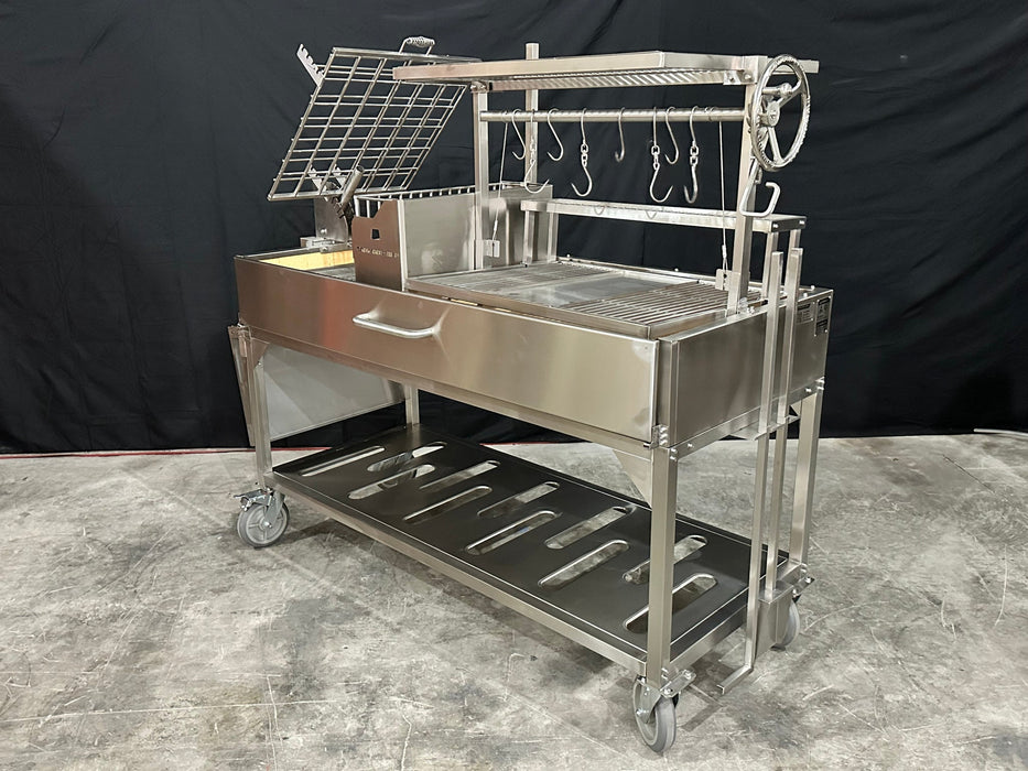 Stono Outdoor Living The Tagwood BBQ Argentine Santa Maria Wood Fire & Charcoal Grill (BBQ26SS) by Tagwood BBQ is a large 304 stainless steel grill with a grate, meat hooks, hand crank, storage shelf, and wheels, shown on a concrete floor against a black background.