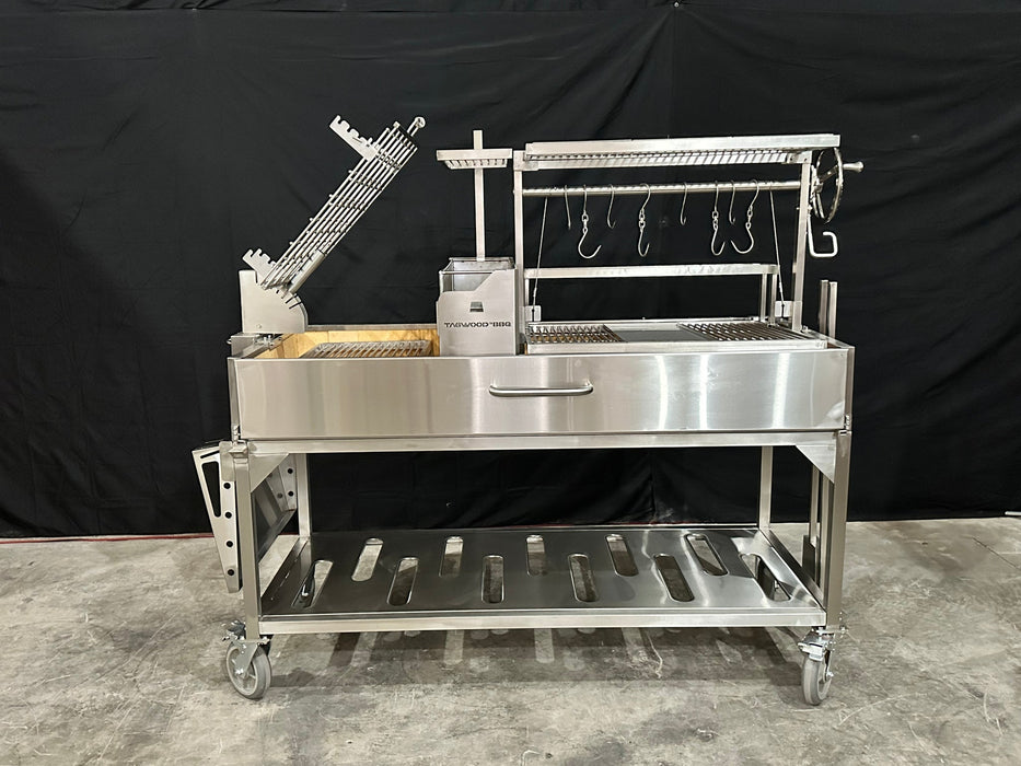 Stono Outdoor Living The Tagwood BBQ Argentine Santa Maria Wood Fire & Charcoal Grill (BBQ26SS) features 304 stainless steel, a side firebox, adjustable grilling racks, hanging hooks, lower storage shelf, and wheels for mobility; shown on concrete with a black backdrop.