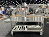 Stono Outdoor Living The Tagwood BBQ Argentine Santa Maria Wood Fire & Charcoal Grill (BBQ26SS), featuring 304 stainless steel, racks, side table, wheels, and storage shelves, is on display at a trade show with people and exhibition booths in the background.