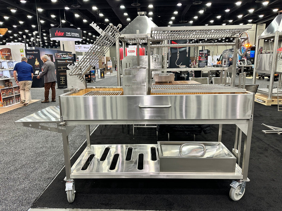 Stono Outdoor Living The Tagwood BBQ Argentine Santa Maria Wood Fire & Charcoal Grill (BBQ26SS), featuring 304 stainless steel, racks, side table, wheels, and storage shelves, is on display at a trade show with people and exhibition booths in the background.