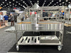Stono Outdoor Living The Tagwood BBQ Argentine Santa Maria Wood Fire & Charcoal Grill (BBQ26SS), a large 304 stainless steel model with shelves and racks, is showcased at a trade show with people and other equipment in the background.