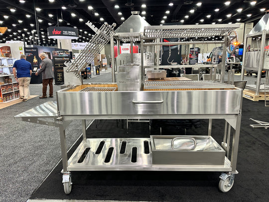 Stono Outdoor Living The Tagwood BBQ Argentine Santa Maria Wood Fire & Charcoal Grill (BBQ26SS), a large 304 stainless steel model with shelves and racks, is showcased at a trade show with people and other equipment in the background.