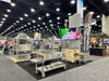 Stono Outdoor Living At a trade show booth, Tagwood BBQ showcases their Argentine Santa Maria Wood Fire & Charcoal Grill (BBQ26SS) with bright lights, banners, and a large screen showing meat prep; people and other booths can be seen in the background.