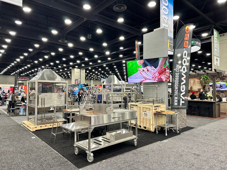 Stono Outdoor Living At a trade show booth, Tagwood BBQ showcases their Argentine Santa Maria Wood Fire & Charcoal Grill (BBQ26SS) with bright lights, banners, and a large screen showing meat prep; people and other booths can be seen in the background.