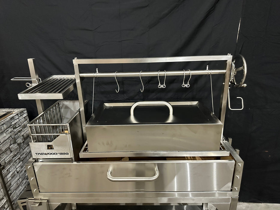 Stono Outdoor Living The Tagwood BBQ Large Top Lid - BBQ59SS by Tagwood BBQ features a stainless steel build, large oven-style lid, rectangular cooking area with main grate, hanging hooks, side shelves, and a large hand crank, shown against a black backdrop.