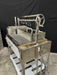 Stono Outdoor Living The Tagwood BBQ Large Top Lid - BBQ59SS by Tagwood BBQ is a stainless steel outdoor grill featuring a spacious grate, side tray, hanging hooks, hand crank, storage compartments, and large top lid for oven-style cooking—shown on a concrete floor with a black backdrop.