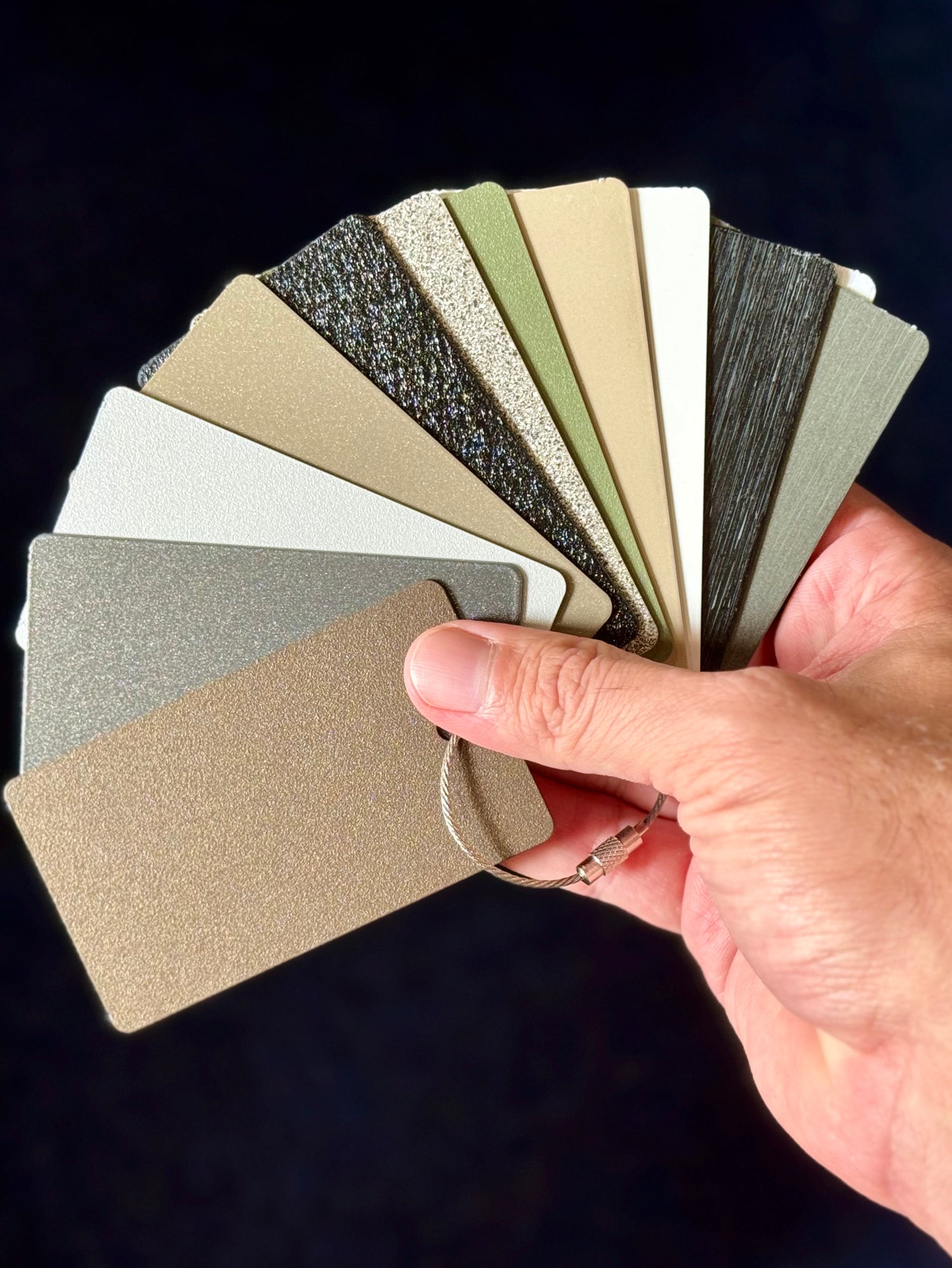Stono Outdoor Living A hand holds a fanned-out selection of textured color sample cards in shades of gray, beige, green, and black against a dark background.