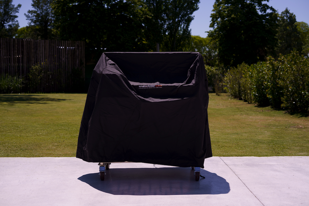 Stono Outdoor Living A Tagwood BBQ Grill Cover for BBQ23SS Grill - BBQ86SS by Tagwood BBQ sits on a wheeled grill on a concrete patio, with green grass and trees behind it on a sunny day.
