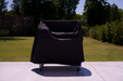Stono Outdoor Living A Tagwood BBQ Grill Cover for BBQ23SS Grill - BBQ86SS by Tagwood BBQ sits on a wheeled grill on a concrete patio, with green grass and trees behind it on a sunny day.