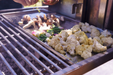 Stono Outdoor Living Fresh cauliflower, mushrooms, and green onions are being grilled on the Tagwood BBQ Interchangeable Griddle for BBQ03SS/05SS - BBQ58SS as a hand arranges the veggies across the surface, with smoke and sunlight enhancing the vibrant scene.