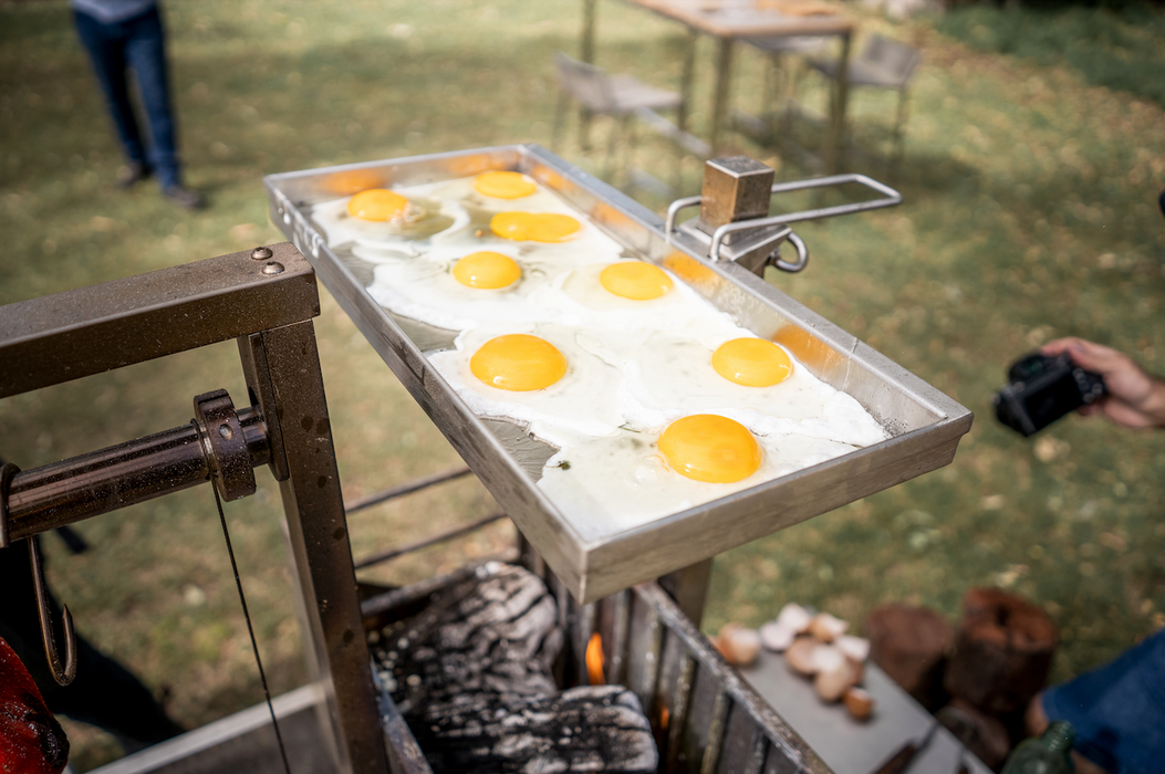 Stono Outdoor Living The Tagwood BBQ Height Adjustable Griddle - BBQ54SS by Tagwood BBQ holds several sunny-side-up eggs sizzling over an outdoor fire, extending the cooking surface with people and equipment visible in the background.
