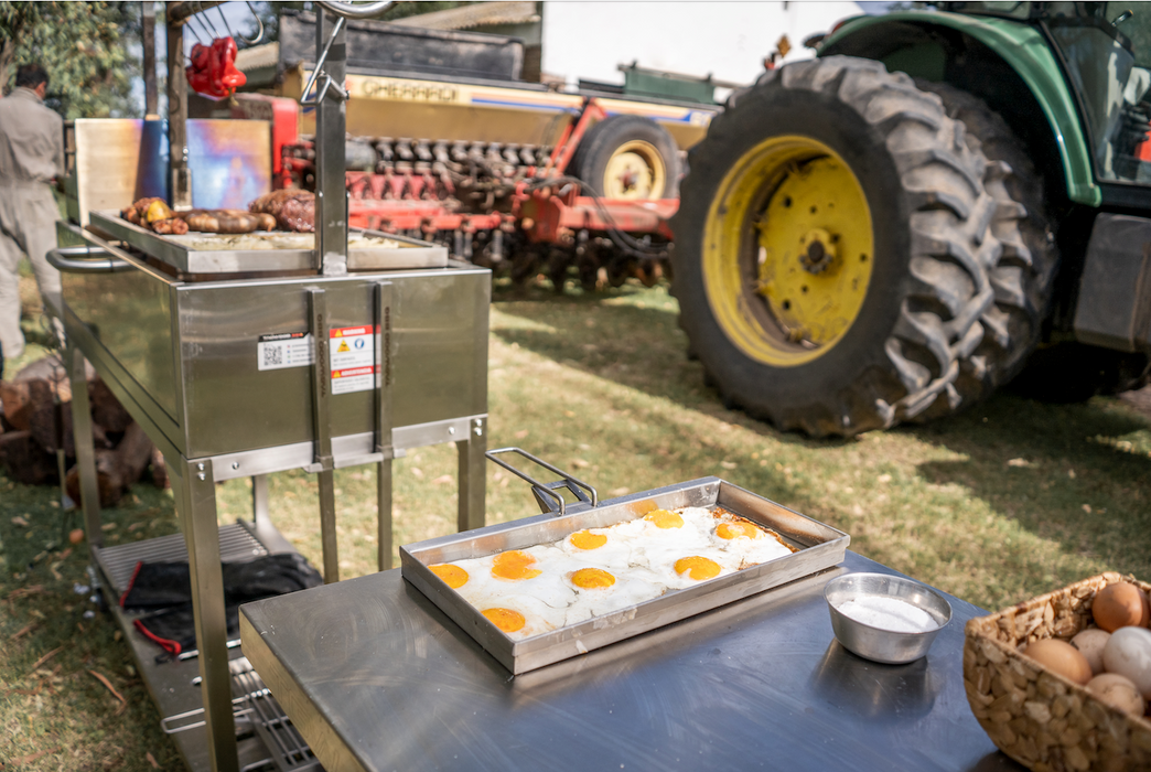 Stono Outdoor Living Outdoors on grass, the Tagwood BBQ Height Adjustable Griddle - BBQ54SS by Tagwood BBQ, made from durable 304 stainless steel, extends cooking space with sunny-side-up eggs on a tray and a bowl of eggs; a tractor sits in the background.