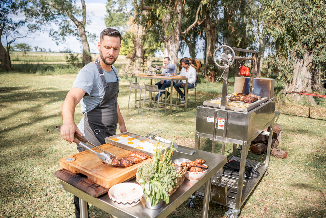Stono Outdoor Living A man in an apron cooks meat and fresh vegetables on the Tagwood BBQ Height Adjustable Griddle - BBQ54SS with a 304 stainless steel surface, while people enjoy a meal under trees in a sunlit, grassy park.